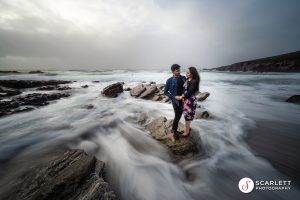 Couple standing on a rock at the beach, a wave has just broken and surrounds them