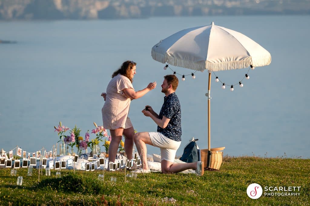 Romantic picnic proposal, Henry is on one knee proposing to Kasia on the clifftops on a summers evening overlooking the sea.