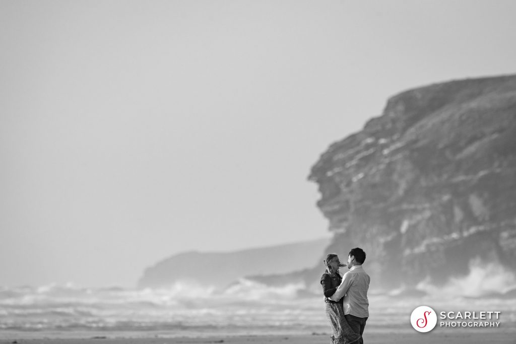 A black and white photo of a couple embracing after accepting a wedding proposal at Watergate Bay with the cliffs and waves in the background.