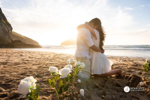 Couple embrace on the beach with the sunset behind them surrounded by roses